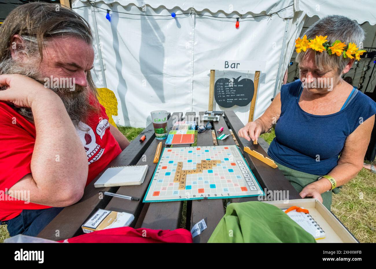 Uomo e donna che indossano un semplice vestito da testa fiorito che suona scrabble su una panchina di legno all'esterno di una tenda della birra in un festival musicale. Foto Stock