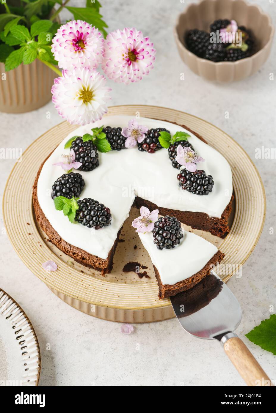 Vista dall'alto. Una fetta di torta al cioccolato fatta in casa con crema alla vaniglia decorata con mora, fiori e foglie di balsamo al limone su una paletta per torta. Foto Stock