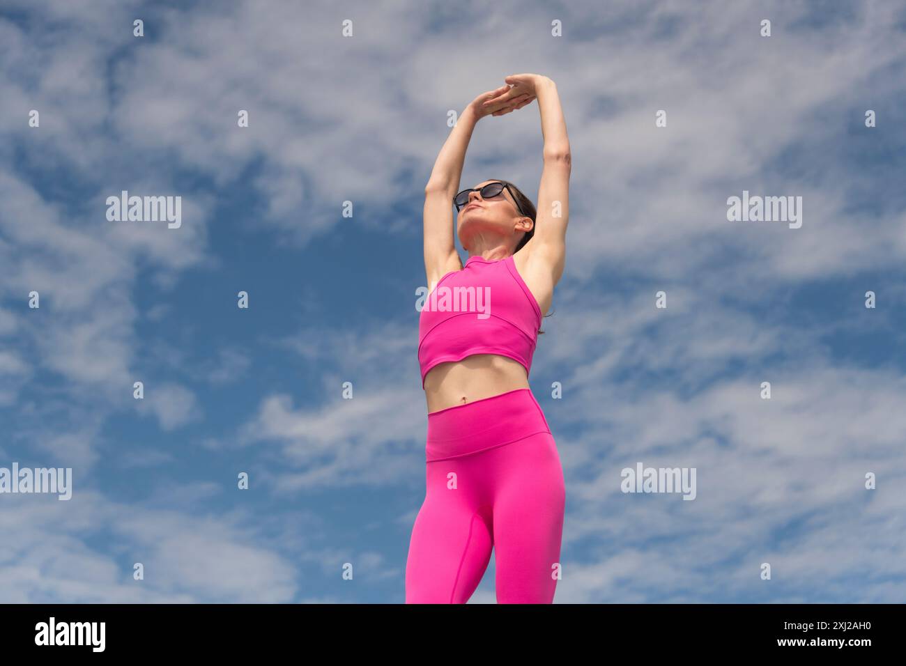 Donna sportiva e in forma che fa esercizi di stretching del braccio indossando abiti sportivi rosa contro il cielo blu Foto Stock