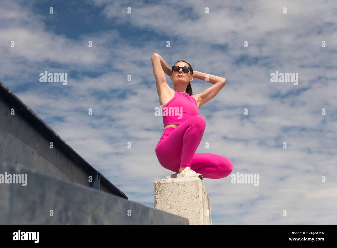 Donna sportiva seduta inginocchiata all'aperto, indossa abiti sportivi rosa, legando i capelli alla schiena prima di correre o allenarsi, sfondo blu del cielo Foto Stock