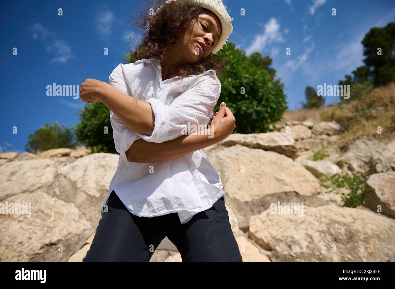 Donna in camicia bianca che posa con sicurezza su uno sfondo roccioso con cielo blu, catturando l'essenza della libertà e della sicurezza di sé. Foto Stock