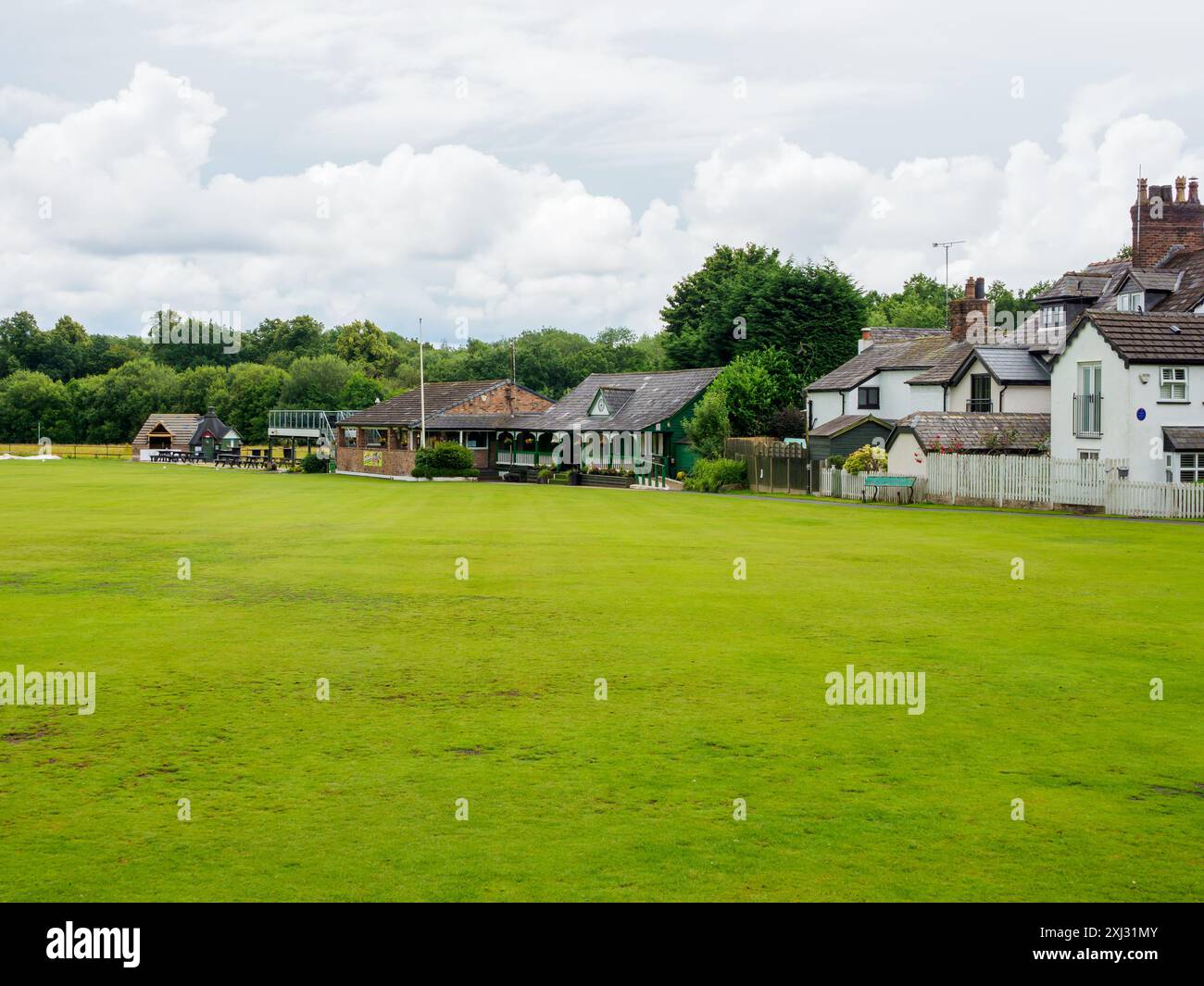 Tranquillo campo da cricket del villaggio circondato da pittoreschi cottage e lussureggiante paesaggio verde sotto un cielo nuvoloso. Foto Stock