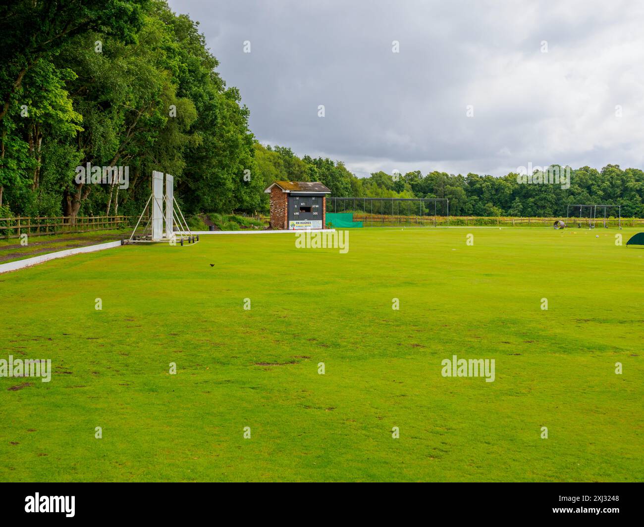 Un lussureggiante campo da cricket verde al Roe Green con tabellone segnapunti e padiglione in una giornata nuvolosa. Foto Stock