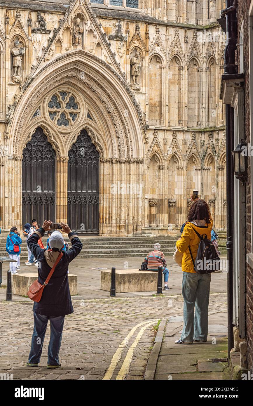 Una vista della Cattedrale di York e della sua intricata architettura gotica con grande porta doppia. I turisti si mescolano vicino alle porte e due turisti in primo piano Foto Stock