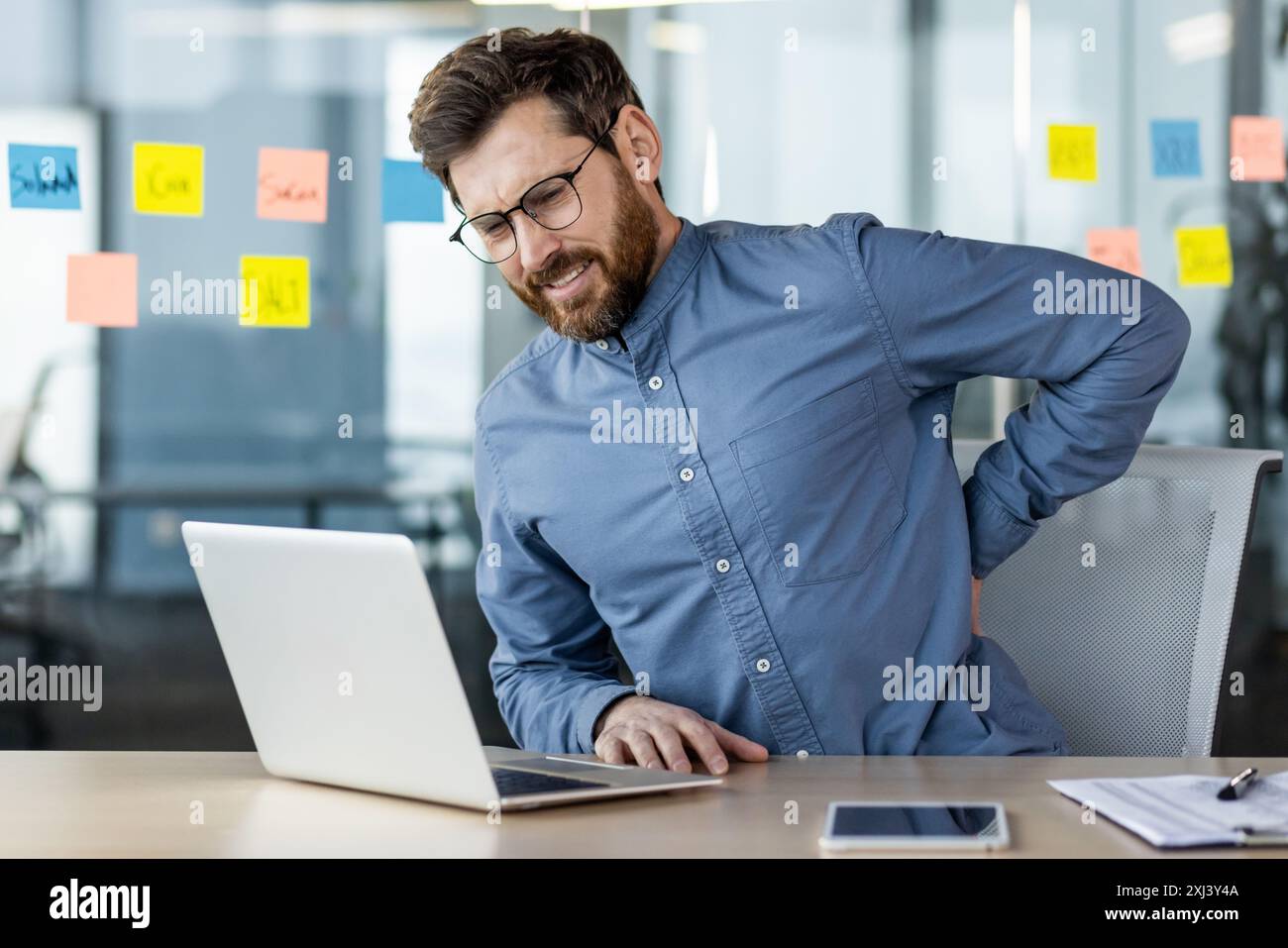 Uomo d'affari con occhiali che soffre di mal di schiena mentre lavora con un computer portatile alla scrivania in un ufficio moderno. Concetto di ergonomia, lavoro in ufficio e disagio da sedute prolungate. Foto Stock