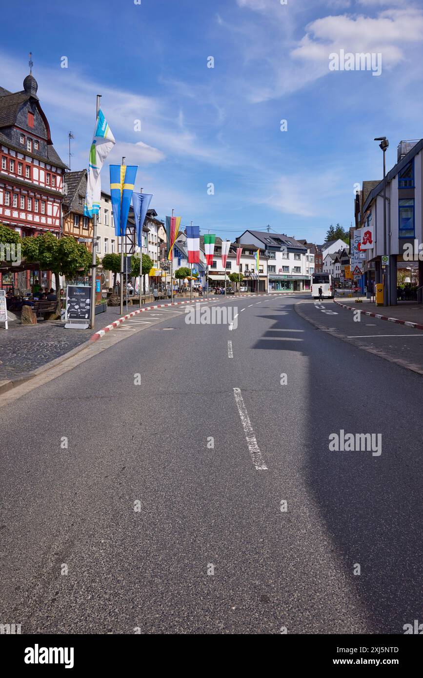 Strada principale con fermata dell'autobus Adenau Markt, edifici, case a graticcio, bandiere e alberi sotto il cielo blu con nuvole di cirrus ad Adenau, Eifel Foto Stock