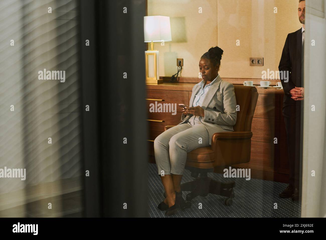 Giovane direttrice finanziaria afroamericana o amministratore delegato in tuta formale seduta in poltrona in sala riunioni e messaggi di testo Foto Stock