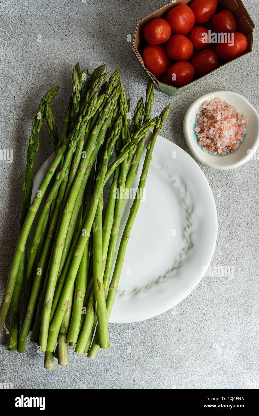 Vista dall'alto di un piatto di asparagi verdi con una scatola di pomodori ciliegini e un piatto di sale rosa dell'himalaya su un tavolo Foto Stock