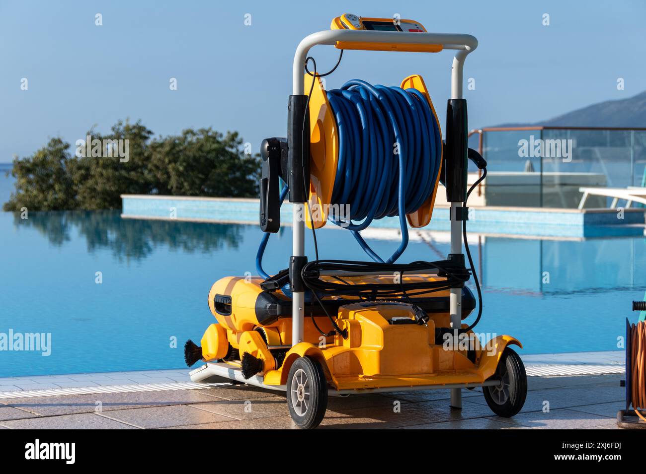 Un pulitore per piscine robotizzato giallo dotato di lunghi tubi a spirale blu, parcheggiato accanto a una tranquilla piscina azzurra che si affaccia sulle tranquille montagne. Avanzate p Foto Stock