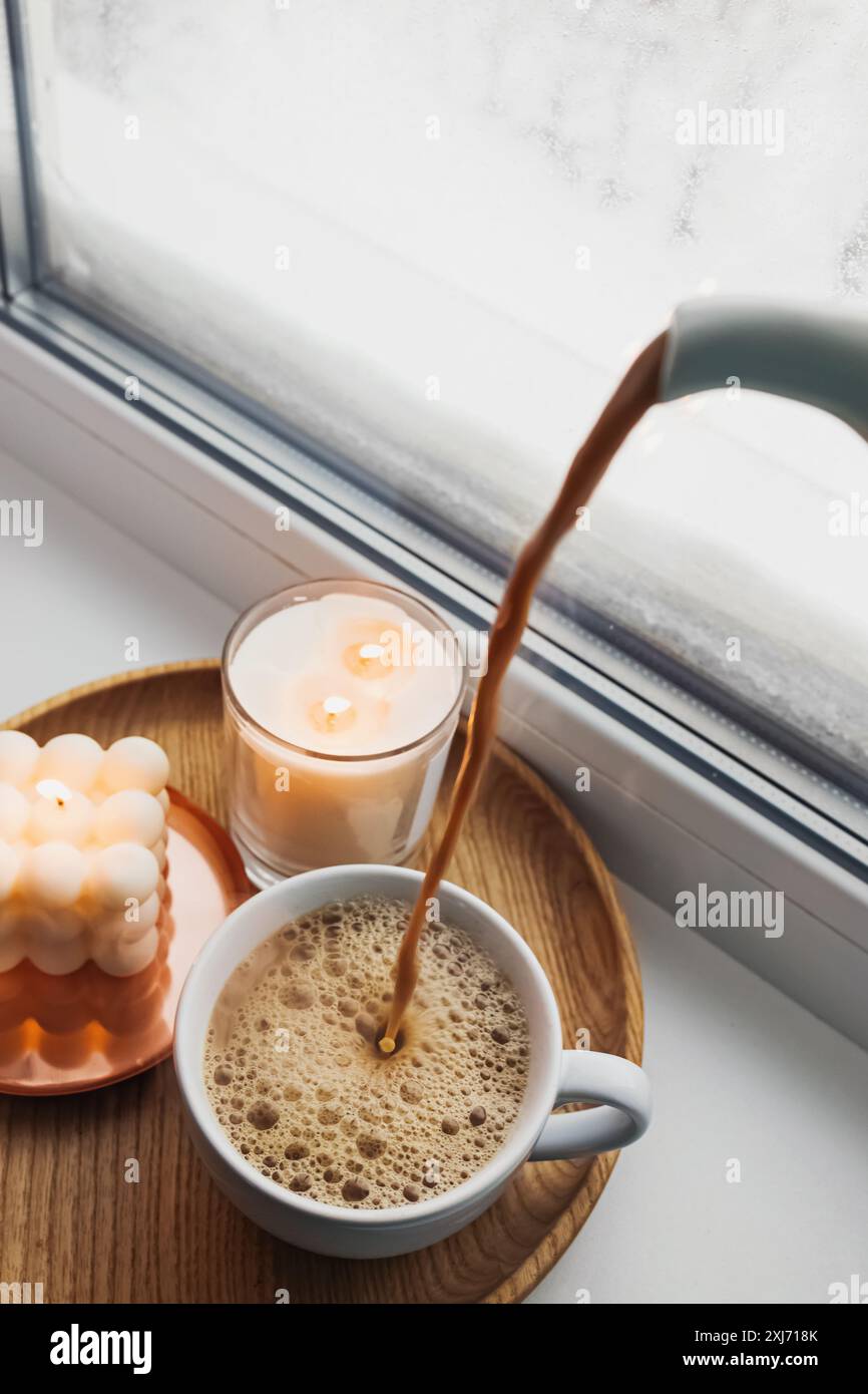 Un ambiente accogliente con caffè versato in una tazza su un vassoio di legno accanto alle candele accese da una finestra ghiacciata. Foto Stock