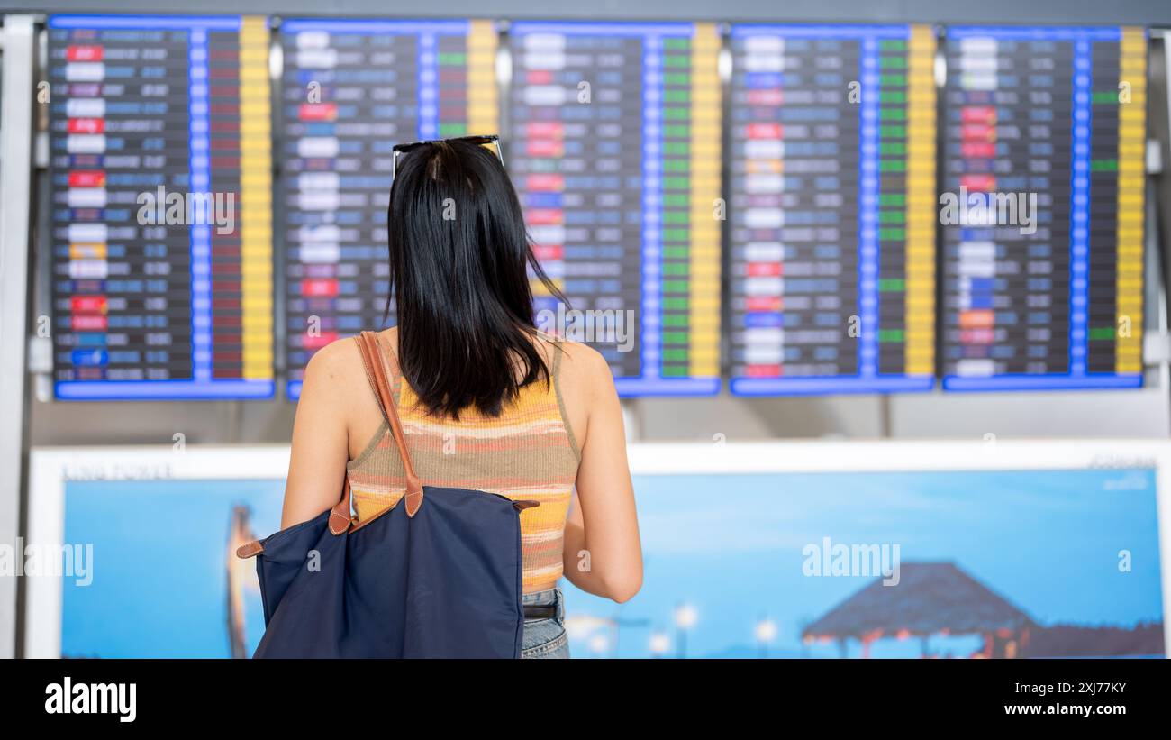 Un'immagine retrovisiva di una donna asiatica che si trova di fronte a una visualizzazione delle informazioni di volo nel terminal dell'aeroporto, controllando l'orario di imbarco. tr Foto Stock