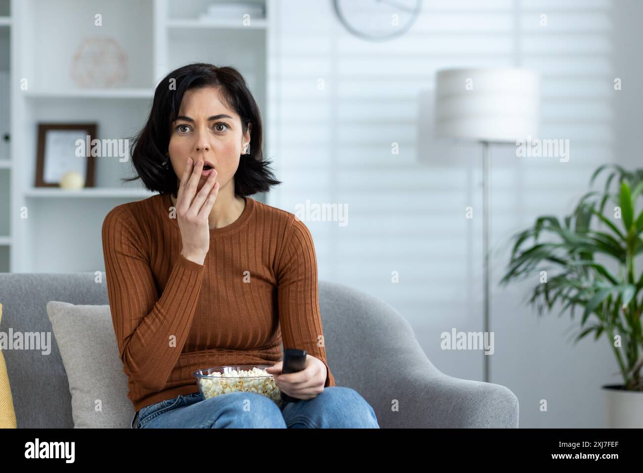 Donna seduta su un divano a casa, che guarda un film horror con un'espressione scioccata. Ha dei popcorn e un telecomando. Foto Stock Donna seduta su un divano a casa, che guarda un film horror con un'espressione scioccata. Ha dei popcorn e un telecomando. Foto Stock