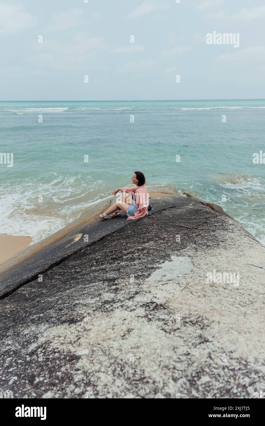 Una donna siede su un grande affioramento roccioso che si affaccia su una spiaggia con acque turchesi. Il cielo è coperto e le onde oceaniche si schiantano dolcemente su t Foto Stock