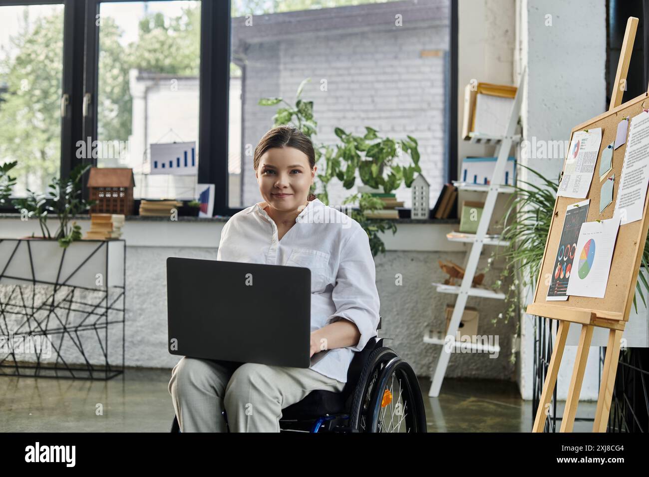 Una giovane donna d'affari con disabilità siede su una sedia a rotelle e lavora su un notebook in un ufficio moderno. Foto Stock