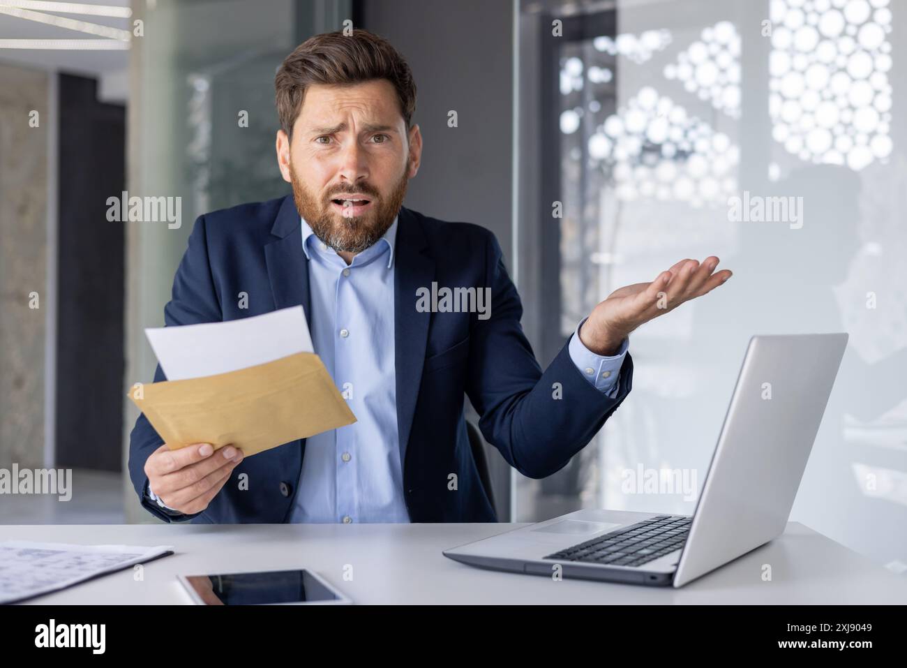 Un uomo d'affari professionista con una tuta blu sembra sconcertato e frustrato mentre esamina un documento alla sua scrivania, ritraendo stress e confusione in un ambiente aziendale. Foto Stock