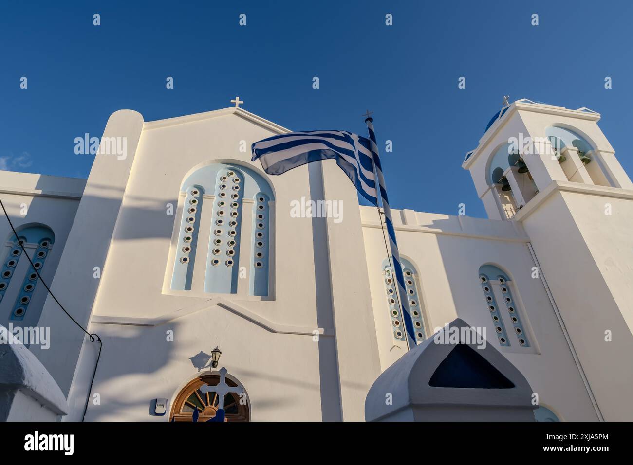 Vista della bandiera greca che sventola nell'aria e di una chiesa imbiancata sullo sfondo a iOS Grecia Foto Stock