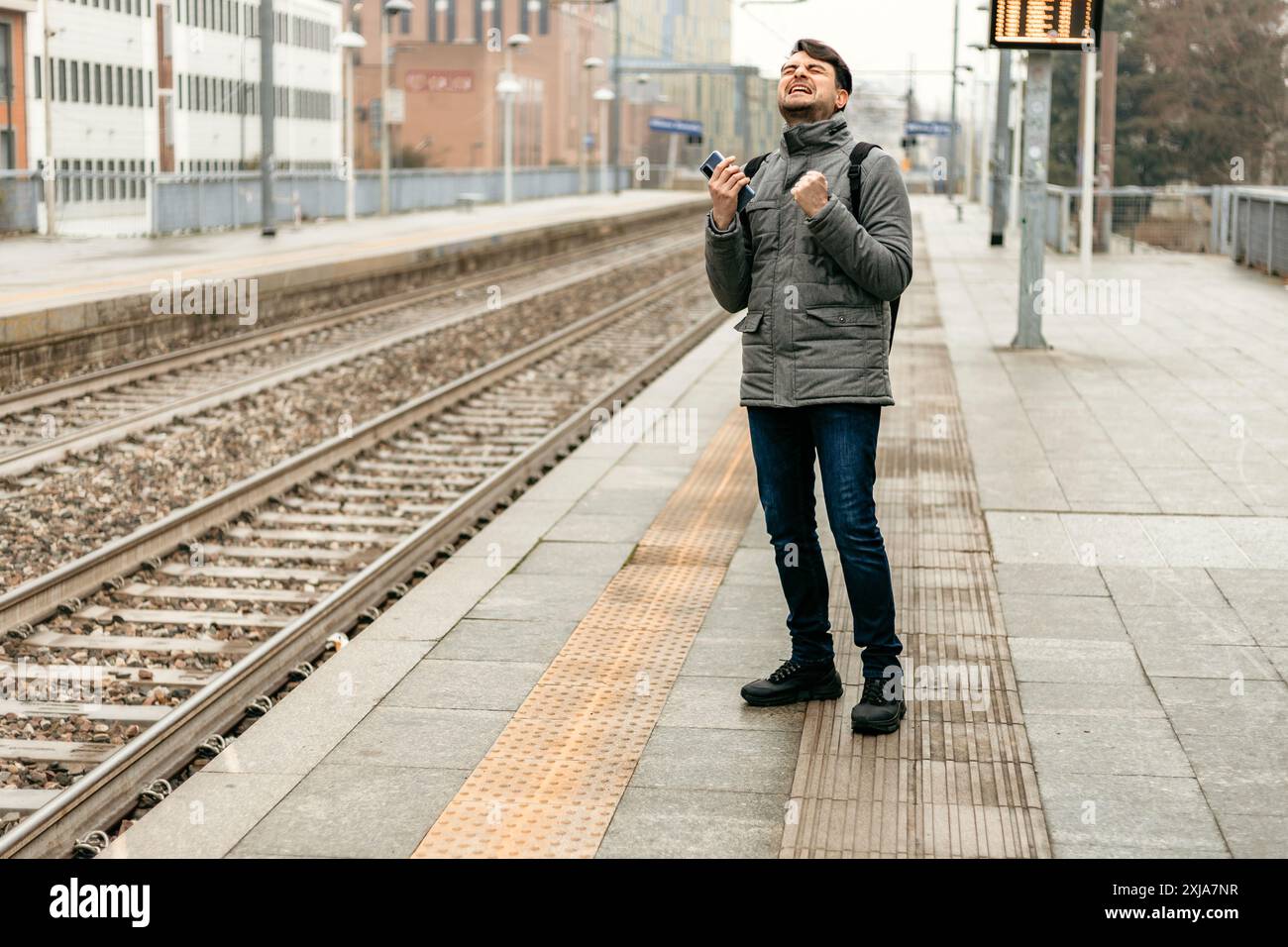 Un uomo con una giacca grigia festeggia con i pugni rialzati su una piattaforma della stazione ferroviaria. Foto Stock