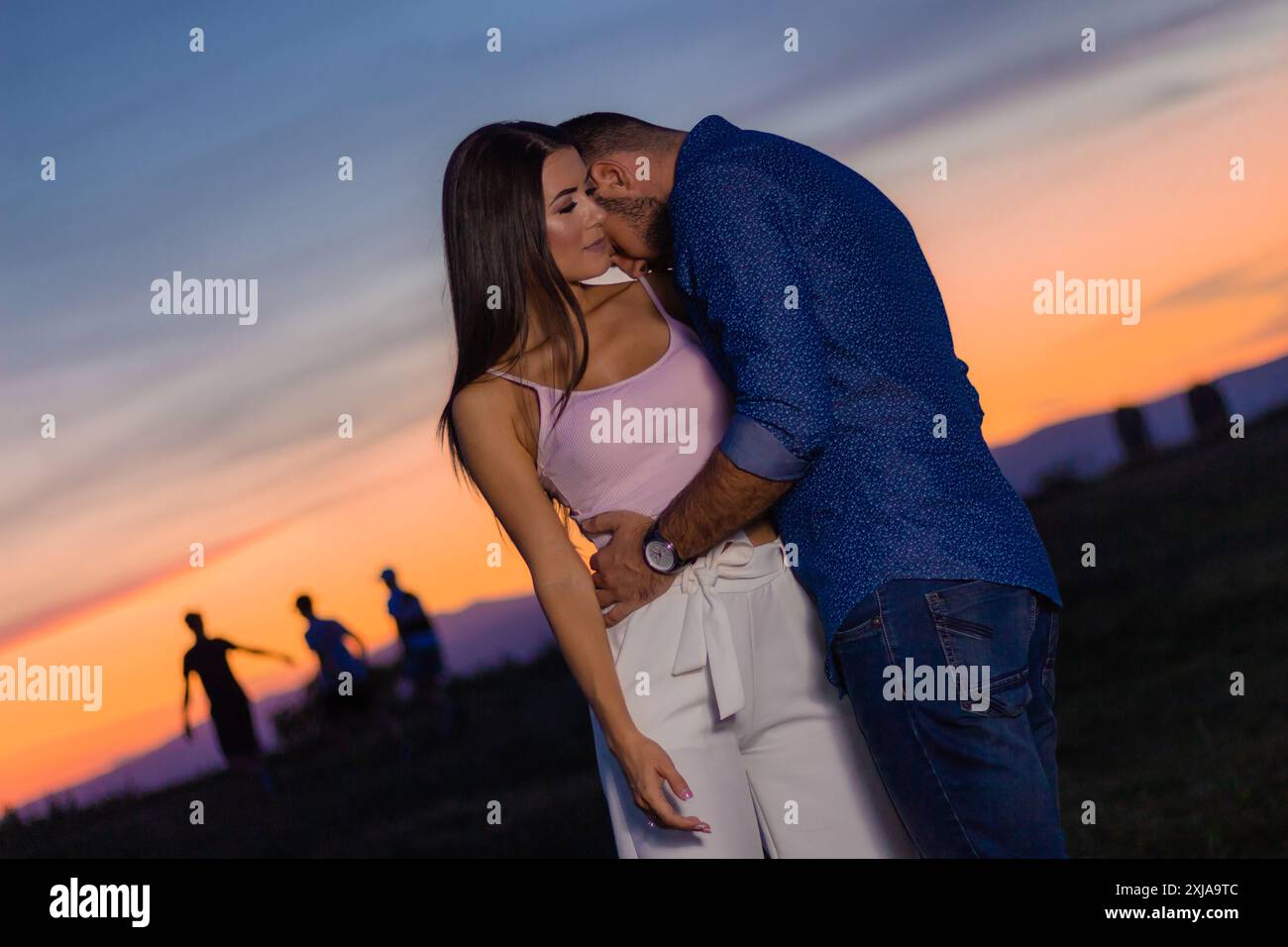 Una coppia abbraccia al tramonto, creando un caldo bagliore dietro di loro. Le sagome delle persone sono visibili in lontananza. Foto Stock