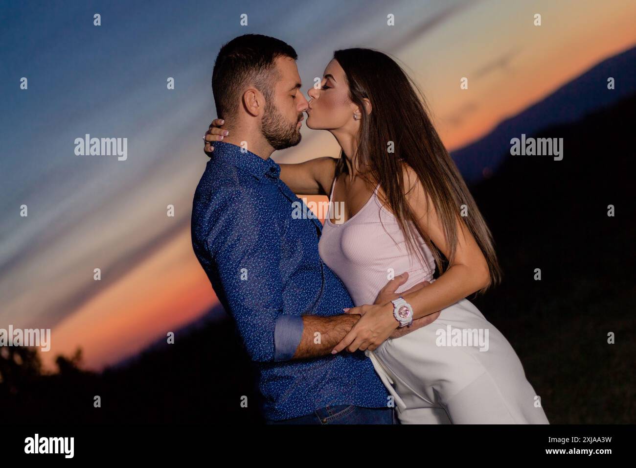 Un uomo e una donna si abbracciano e si baciano mentre sono in piedi in un campo al tramonto. Foto Stock