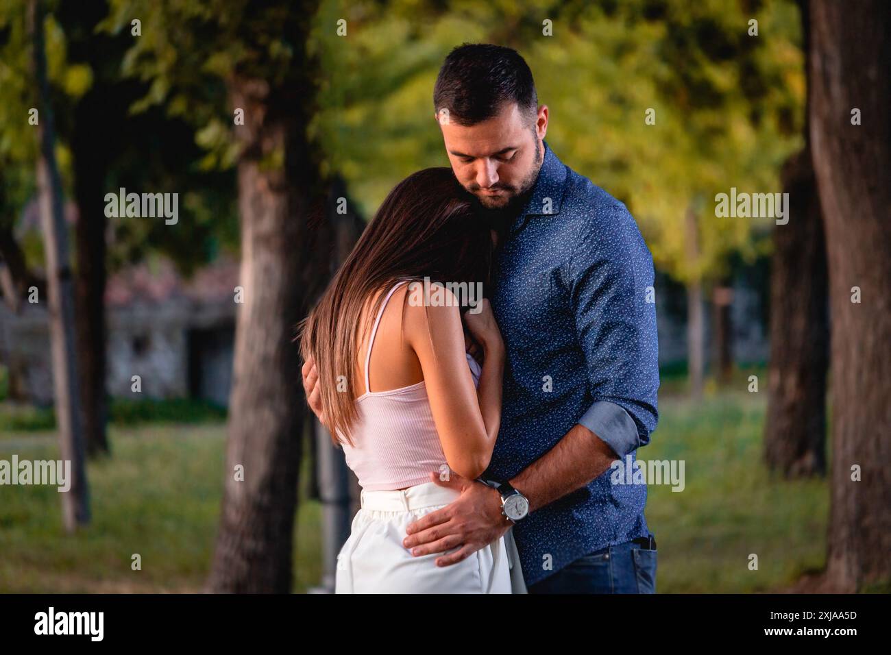 Un uomo con la barba conforta una donna mentre si trova in un parco. La donna indossa una canotta bianca e pantaloni bianchi. L'uomo indossa un bottone blu. Foto Stock