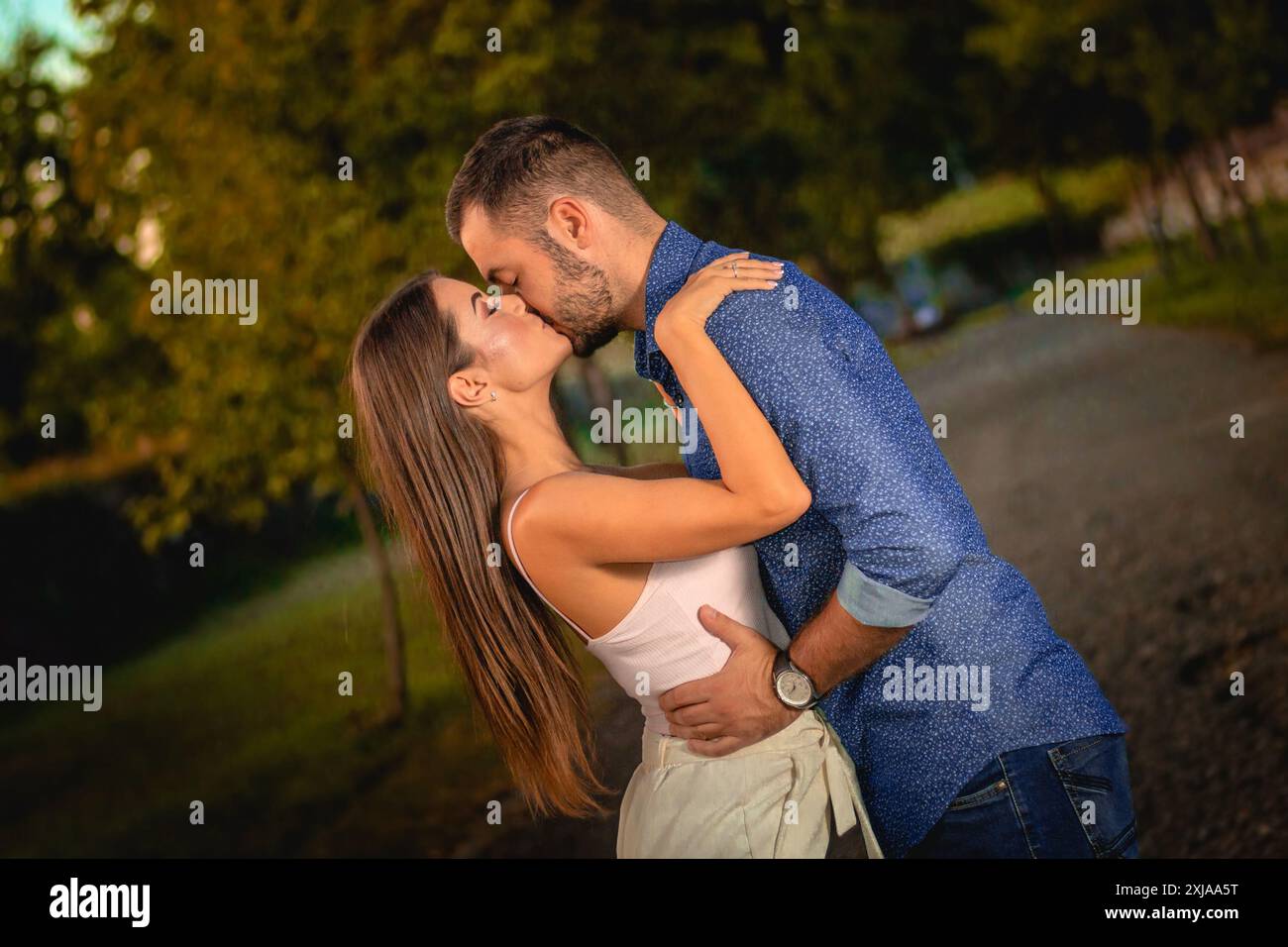 Una giovane coppia sta baciando in un parco durante il tramonto, con alberi sullo sfondo. Foto Stock