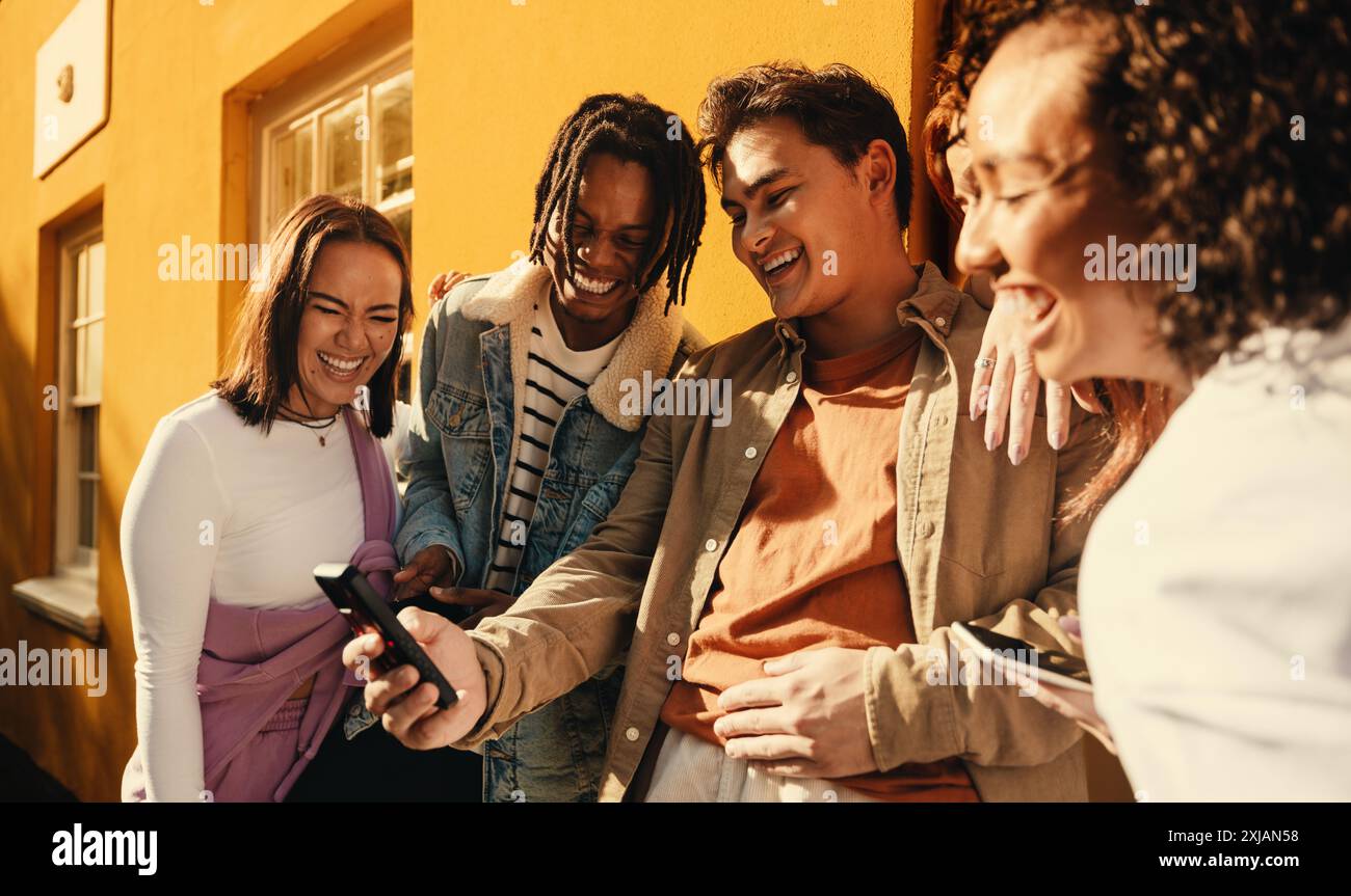 Un gruppo felice di amici si è riunito all'esterno, condividendo un momento divertente mentre guardavano un telefono. Social media e amicizia in un ambiente soleggiato. Foto Stock Un gruppo felice di amici si è riunito all'esterno, condividendo un momento divertente mentre guardavano un telefono. Social media e amicizia in un ambiente soleggiato. Foto Stock