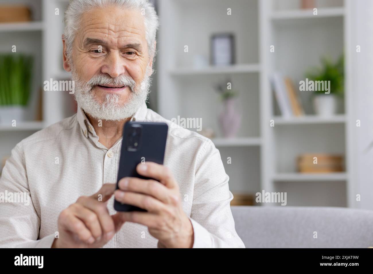 Foto ravvicinata di un nonno anziano sorridente in pensione con una camicia bianca e capelli grigi che è a casa e utilizza uno smartphone. Foto Stock