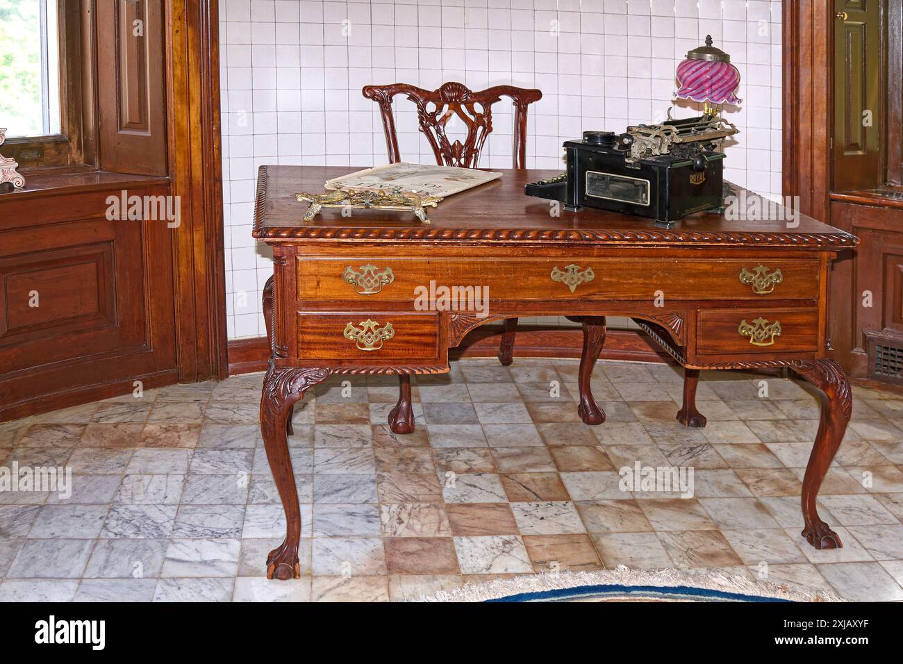scrivania, sedia, vecchia macchina da scrivere, rivista, piccola lampada in vetro, pavimento piastrellato, pareti piastrellate bianche, sedili con finestre in legno Foto Stock