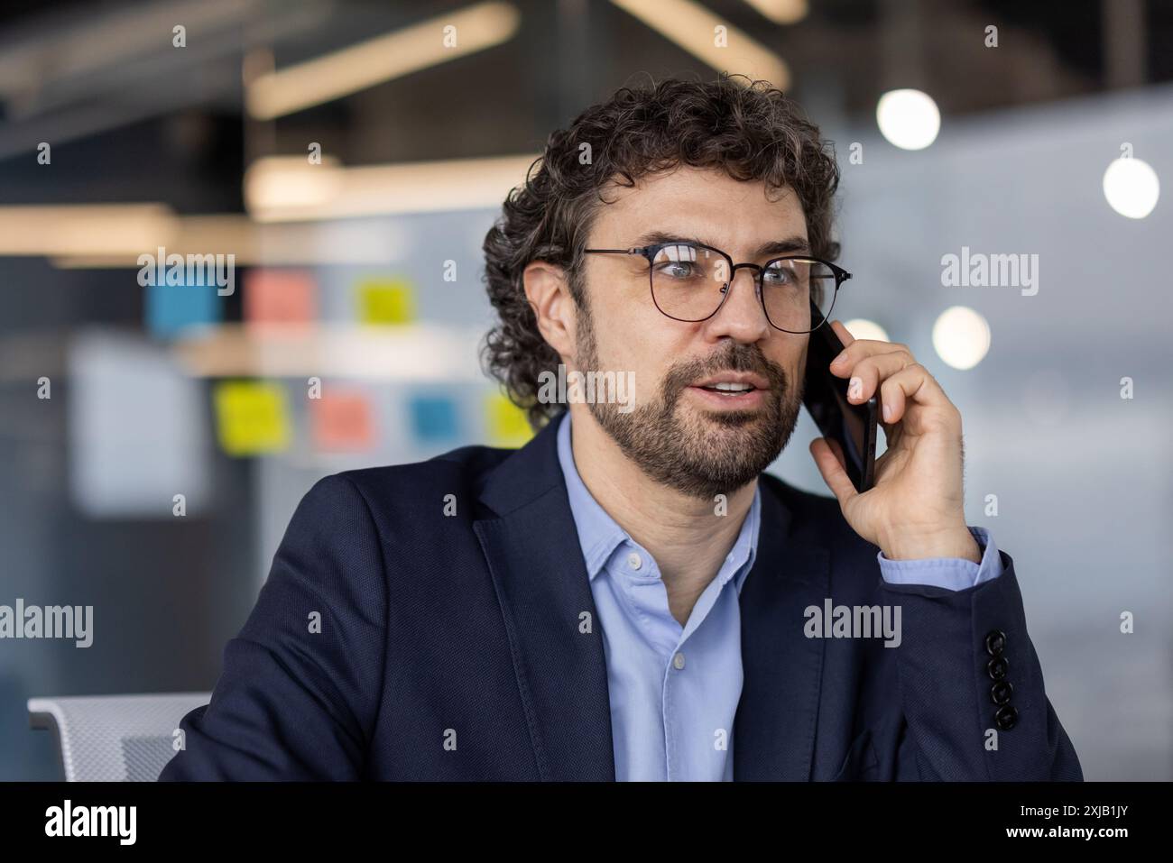 Uomo d'affari in costume blu scuro che parla al telefono in un ufficio moderno, circondato da note adesive colorate e impegnato in una conversazione professionale. SCE Foto Stock