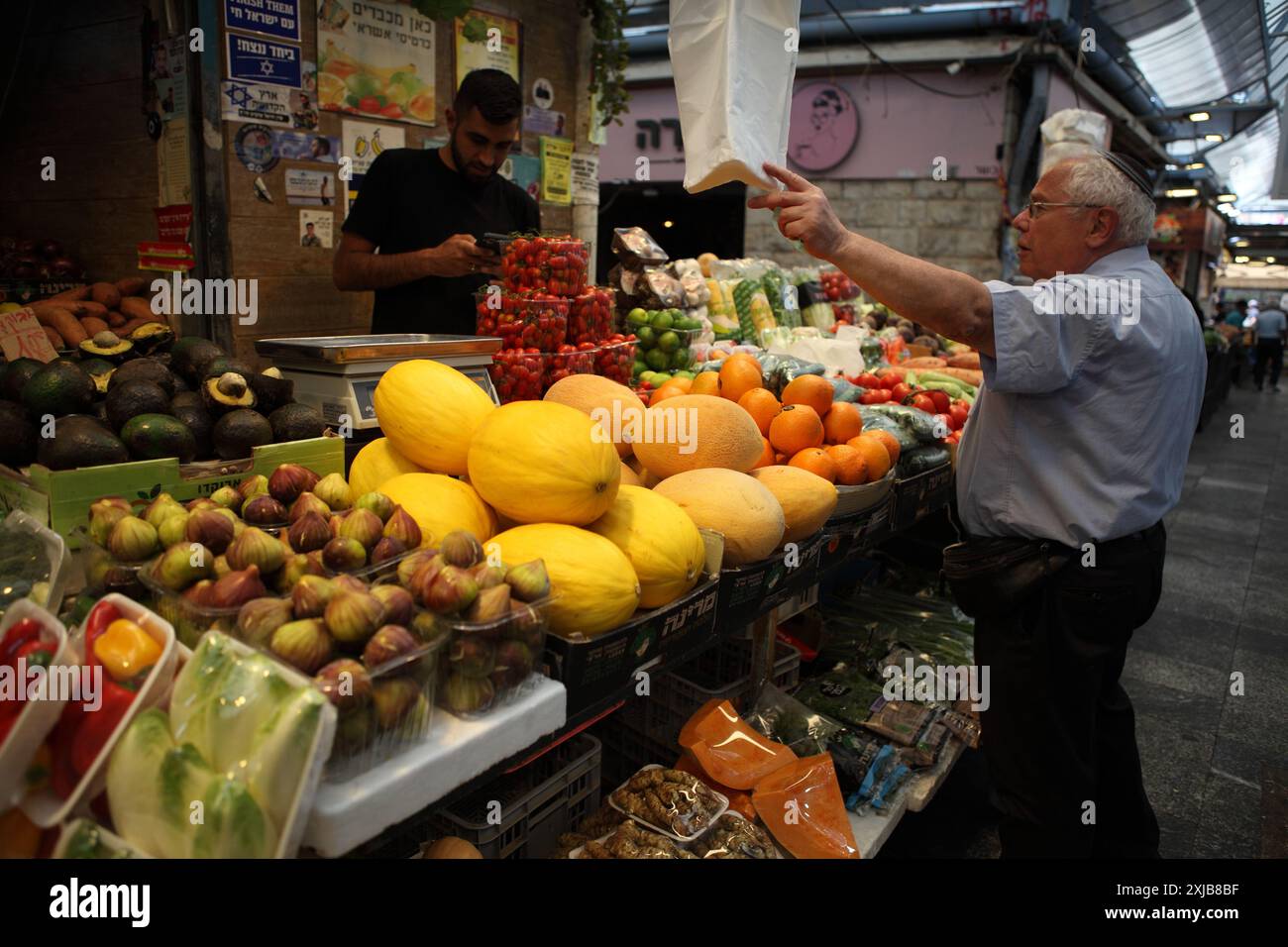 Un uomo israeliano che indossa abiti estivi acquista da un venditore di prodotti in un mercato di Machane Yehuda qui vengono venduti fichi, avocado, melone, arance e pomodori ciliegini Foto Stock
