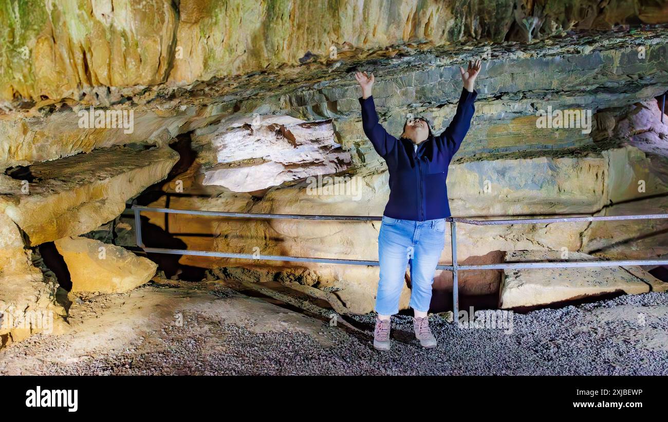 Donna turistica che tocca il soffitto di una grotta all'interno di una formazione rocciosa al Castello di Logne, pareti rocciose, irregolari e scanalate, ringhiere in metallo, abbigliamento casual blu Foto Stock