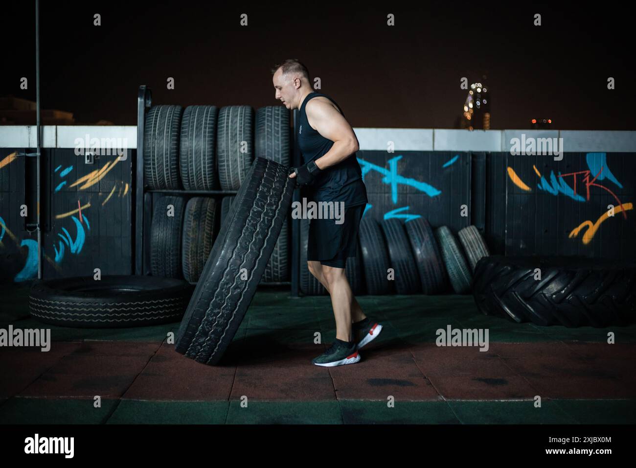 Un uomo con un abbigliamento da ginnastica nero solleva una grande gomma durante un allenamento notturno sul tetto. Foto Stock