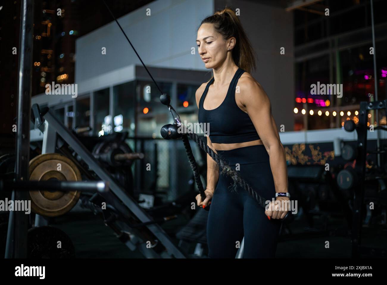 Una donna con abbigliamento atletico nero esegue un esercizio di fila per macchine cablate in palestra, concentrandosi sulla sua forma e coinvolgendo i muscoli della schiena. Foto Stock