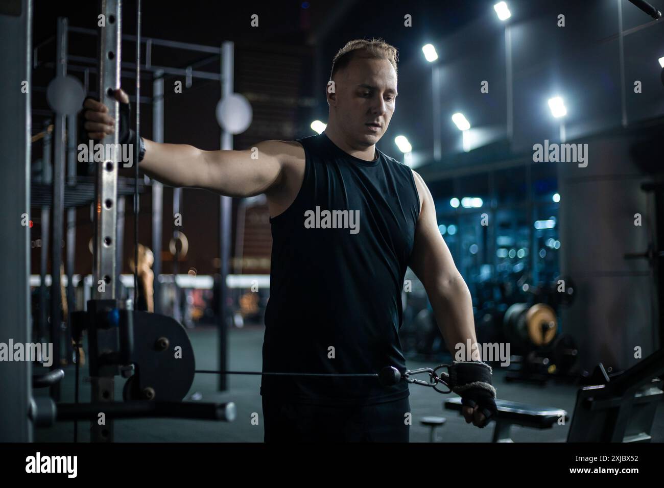 Un uomo con canotta nera esegue un esercizio di pull-down con cavo mentre guarda concentrato in una moderna palestra di sera. Foto Stock