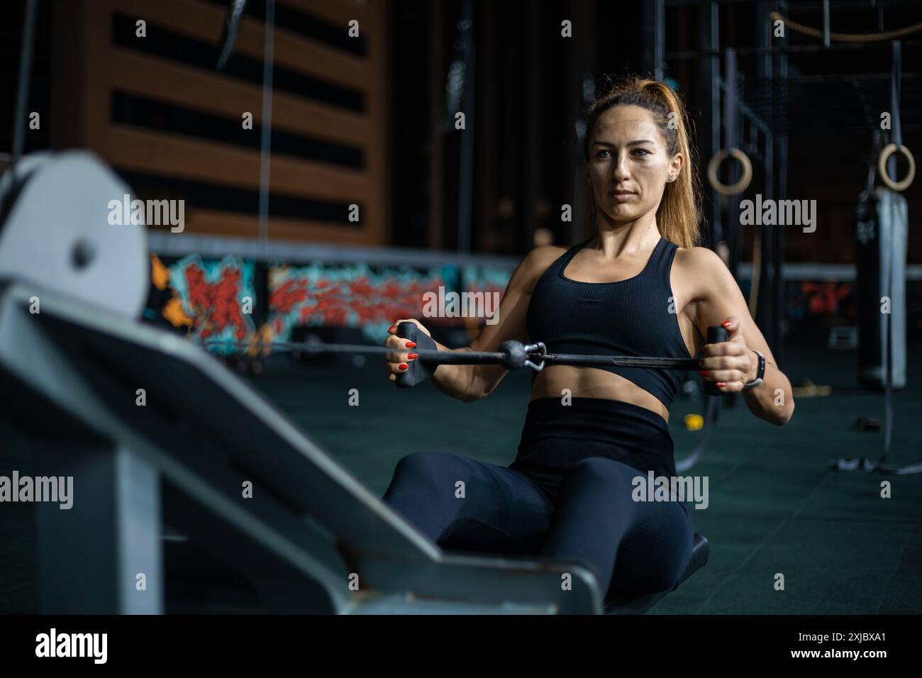 Una donna con abiti da ginnastica neri usa una macchina a remi in una palestra moderna. Foto Stock