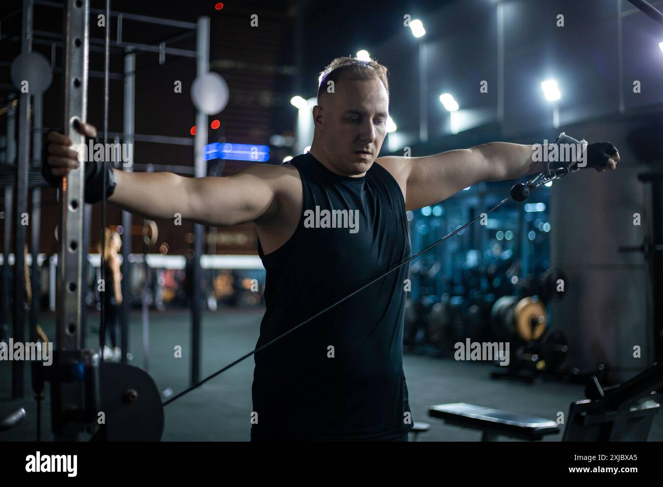 Un uomo con canotta nera si allena con una macchina via cavo in un centro fitness di notte. Foto Stock