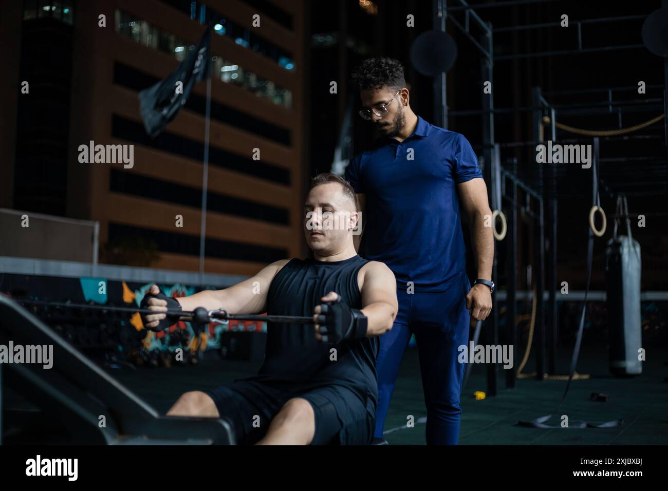 Un uomo usa una vogatrice con un allenatore che guarda da vicino durante un allenamento serale. Foto Stock