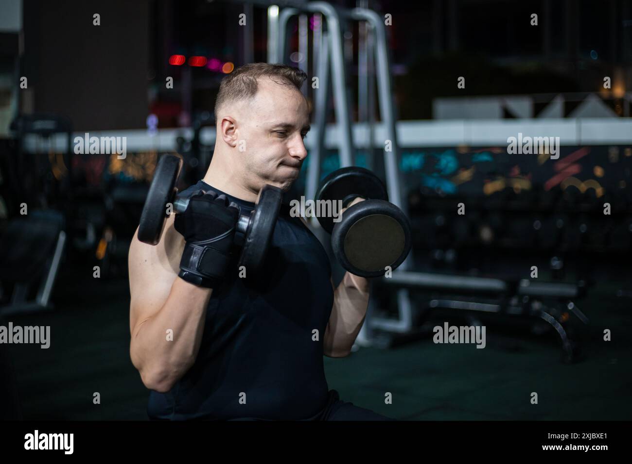 Un uomo con una camicia nera solleva i manubri durante un allenamento serale in palestra. Foto Stock