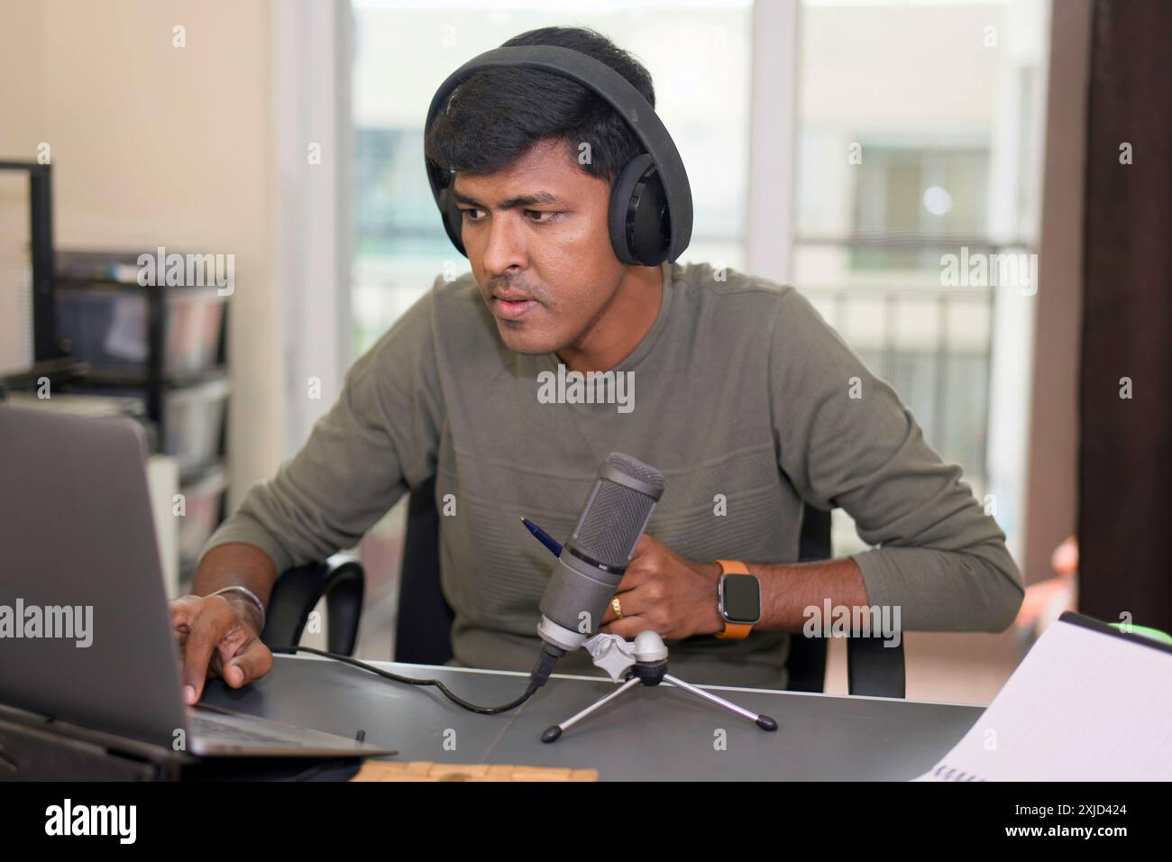 Giovane uomo che registra intensamente un podcast e modifica l'audio sul suo computer portatile con una configurazione professionale del microfono. Foto Stock