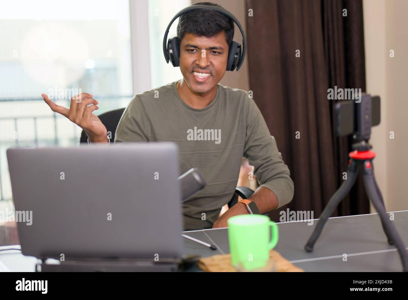 L'uomo sorridente coinvolge gli spettatori con gesti vivaci durante il live streaming domestico, utilizzando un laptop, cuffie e una configurazione per smartphone. Foto Stock