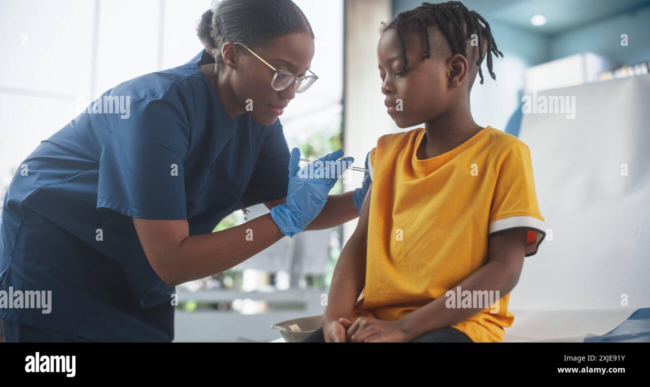 Giovane ragazzo afroamericano seduto sulla sedia al Bright Hospital e ottenere il suo vaccino polio. Infermiera nera donna sta eseguendo l'iniezione. Una donna professionale parla con Desired Kid e lo calma Foto Stock