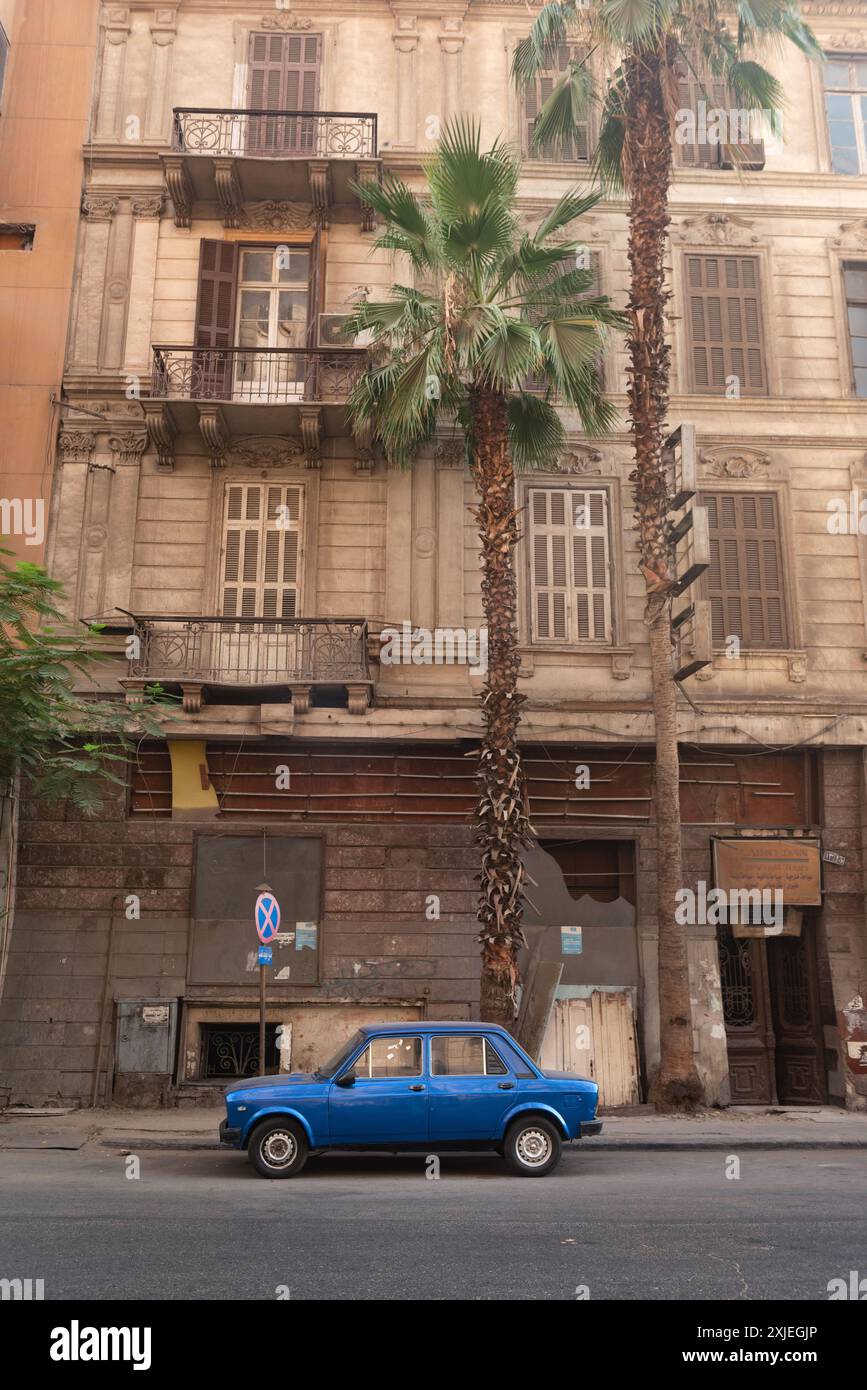 Cairo, Egitto. 1° luglio 2024 Una piccola auto blu parcheggiata in una strada vuota del centro del Cairo, con palme e tipica architettura egiziana Foto Stock