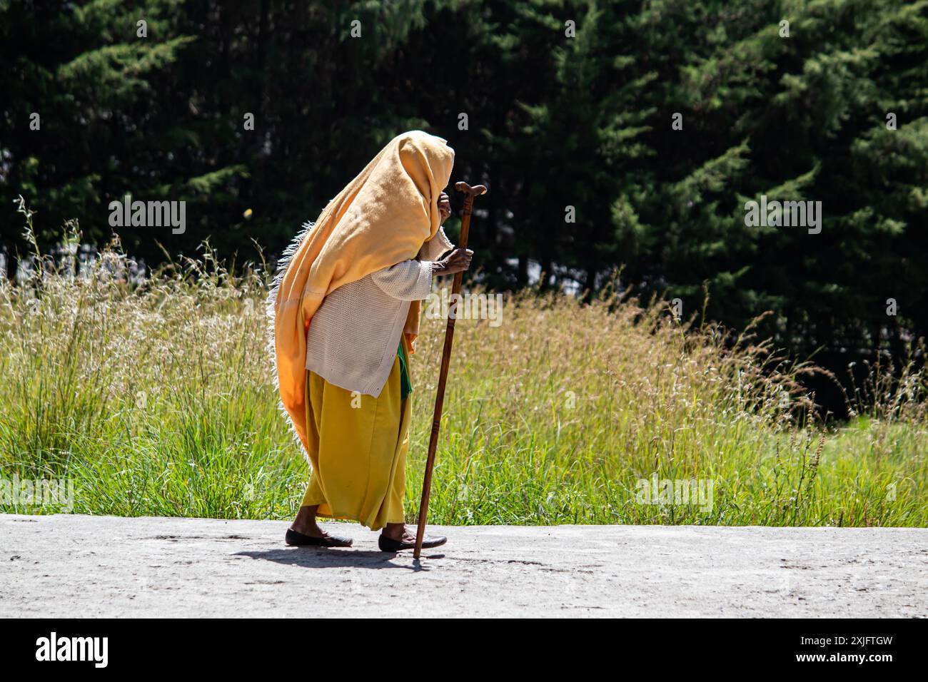 Una vecchia mamma etiope, tradizionalmente vestita di giallo, cammina con un bastone di legno, riflettendo il patrimonio culturale, la dignità e la resilienza Foto Stock