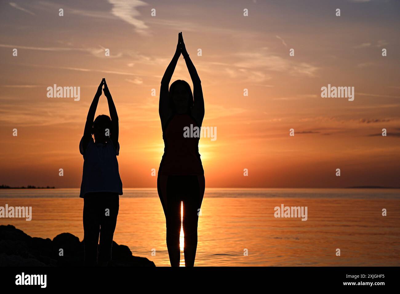 Madre e figlia praticano yoga sulla spiaggia al tramonto, godendosi un momento di pace insieme. Foto Stock