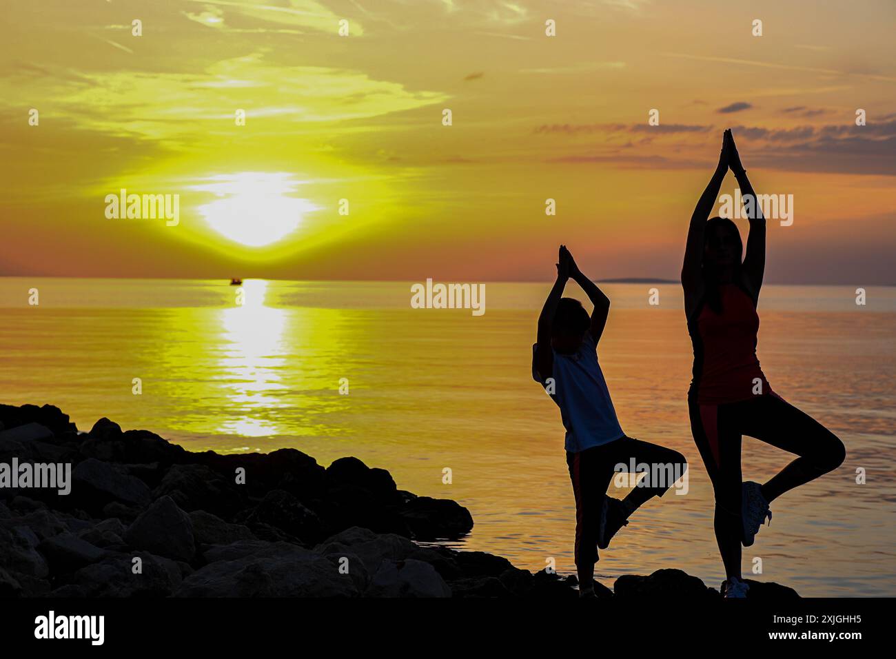 Madre e figlia praticano yoga sulla spiaggia al tramonto, godendosi un momento di pace insieme. Foto Stock