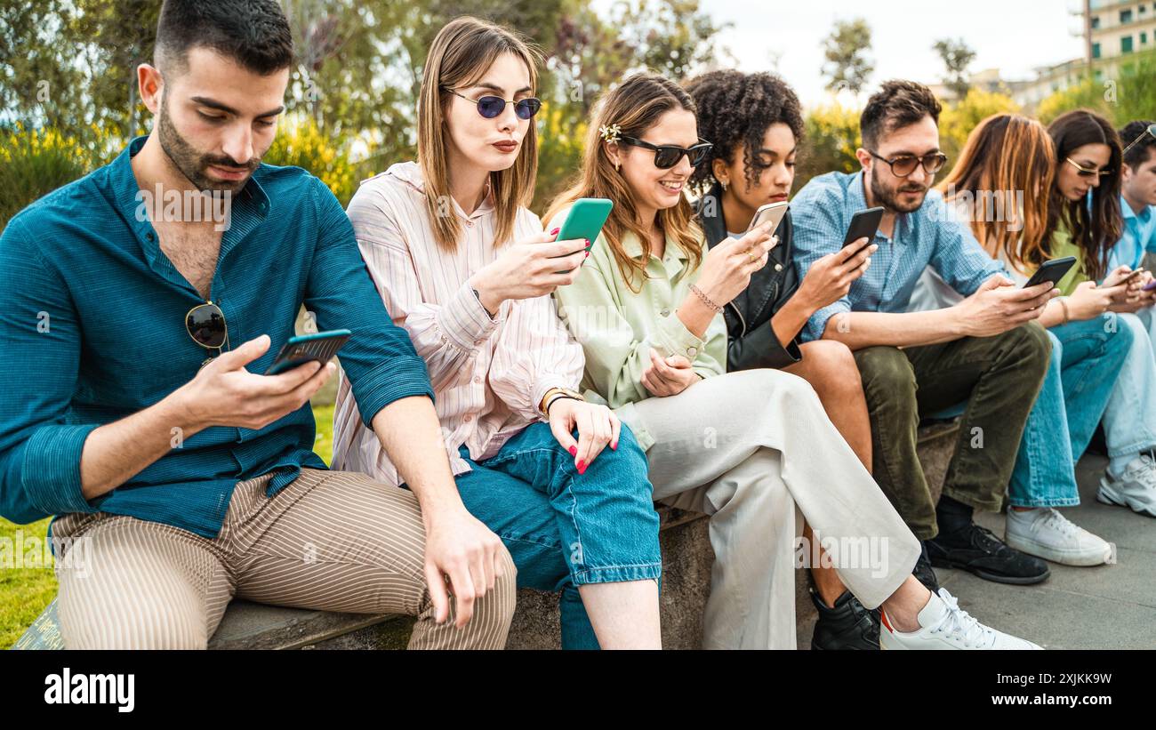 Amici multietnici seduti insieme nel parco urbano, tutti concentrati sui dispositivi mobili. Illustra l'utilizzo dei social media, i nativi digitali e i dipendenti degli smartphone Foto Stock