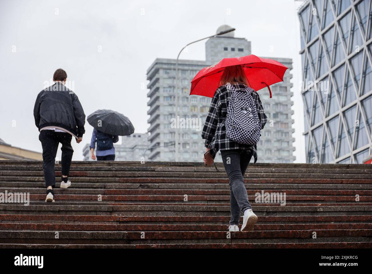 Persone con ombrelloni che camminano su per le scale sullo sfondo degli edifici della città. Pioggia nella città estiva Foto Stock