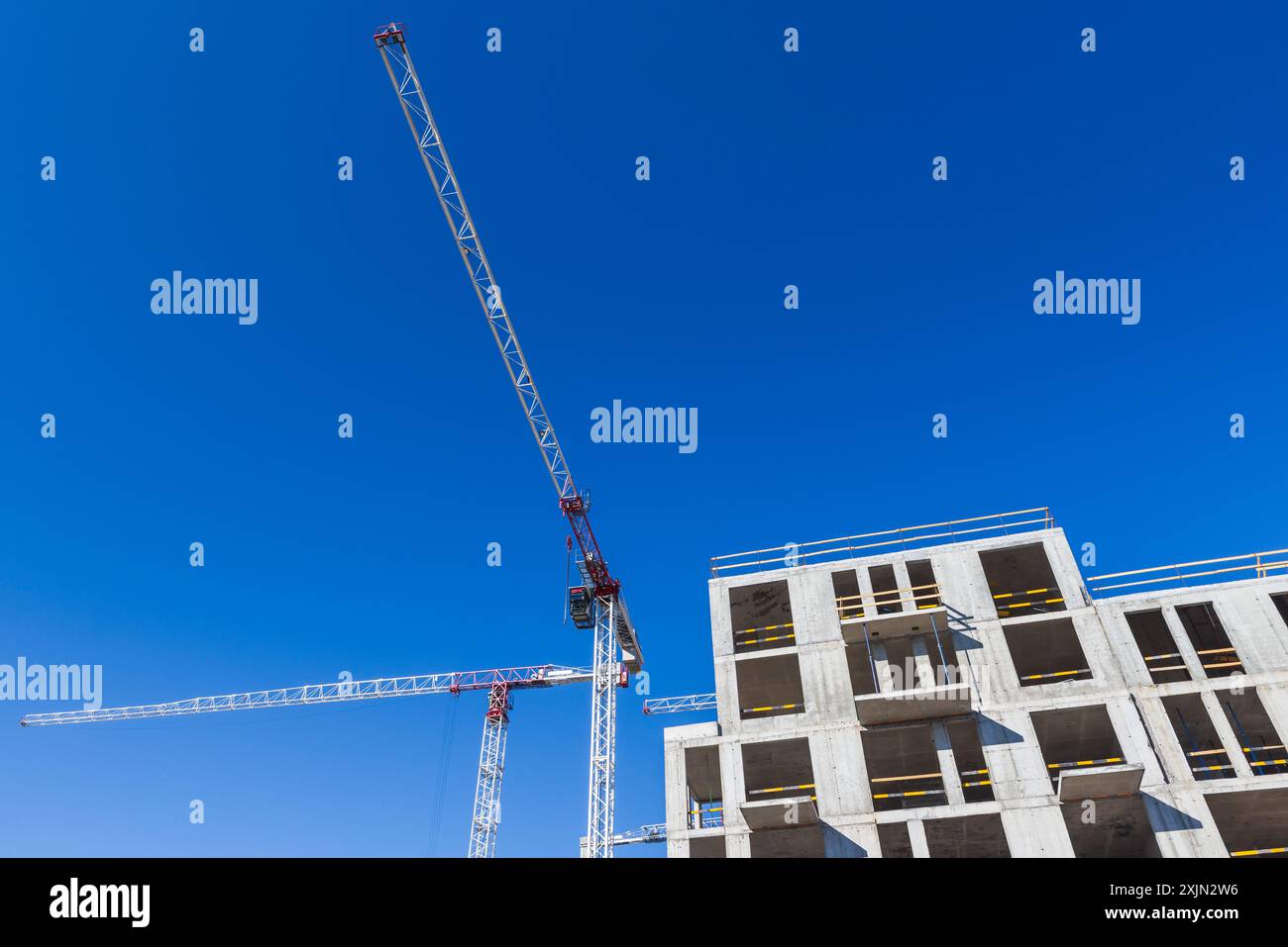 Le gru da costruzione e la moderna carcassa in cemento con finestre vuote sono sotto il cielo blu Foto Stock