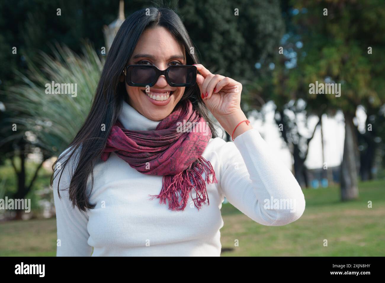 Ritratto di una giovane donna che guarda la macchina fotografica sul bordo dei suoi occhiali, è nel parco della città di Buenos Aires, è felice e sorridente Foto Stock