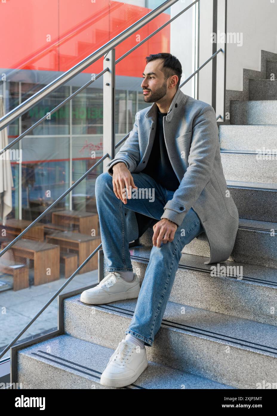 Ritratto di un bel giovane uomo. Un uomo asiatico sorridente con una barba ordinata si siede sui gradini vicino alla finestra e guarda in lontananza Foto Stock